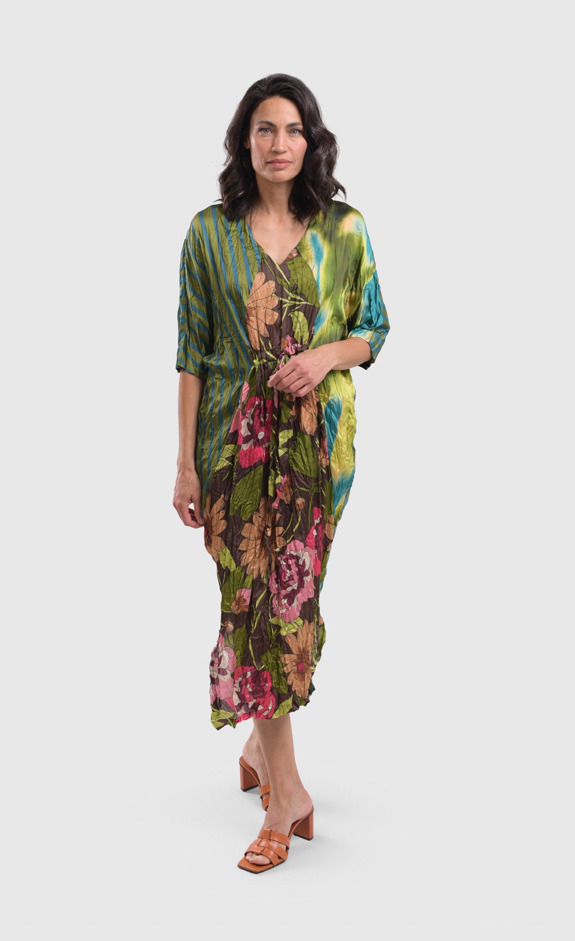 Front full body view of a woman wearing the alembika skye caftan dress in the color aquamarine. This dress has green, yellow, and blue tie dye on the left side, blue and green stripes on the right side and back, and pink flowers on a brown backdrop down the center of the front. The center of the front of the dress is tied to bring the waist in.