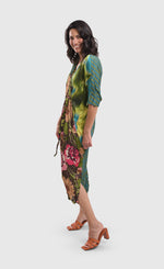 Load image into Gallery viewer, Left side full body view of a woman wearing the alembika skye caftan dress in the color aquamarine. This dress has green, yellow, and blue tie dye on the left side, blue and green stripes on the back, and pink flowers on a brown backdrop down the center of the front. The center of the front of the dress is tied to bring the waist in.
