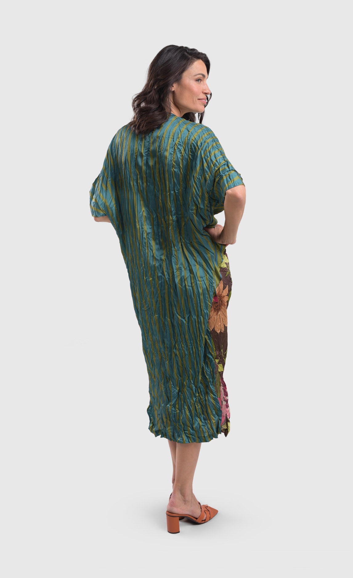 Back full body view of a woman wearing the alembika skye caftan dress in the color aquamarine. This dress has blue and green stripes on the right side and back. The dress sits below the knees.