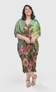 Front full body view of a woman wearing the alembika skye caftan dress in the color aquamarine. This dress has green, yellow, and blue tie dye on the left side, blue and green stripes on the right side and back, and pink flowers on a brown backdrop down the center of the front. The center of the front of the dress is tied to bring the waist in.