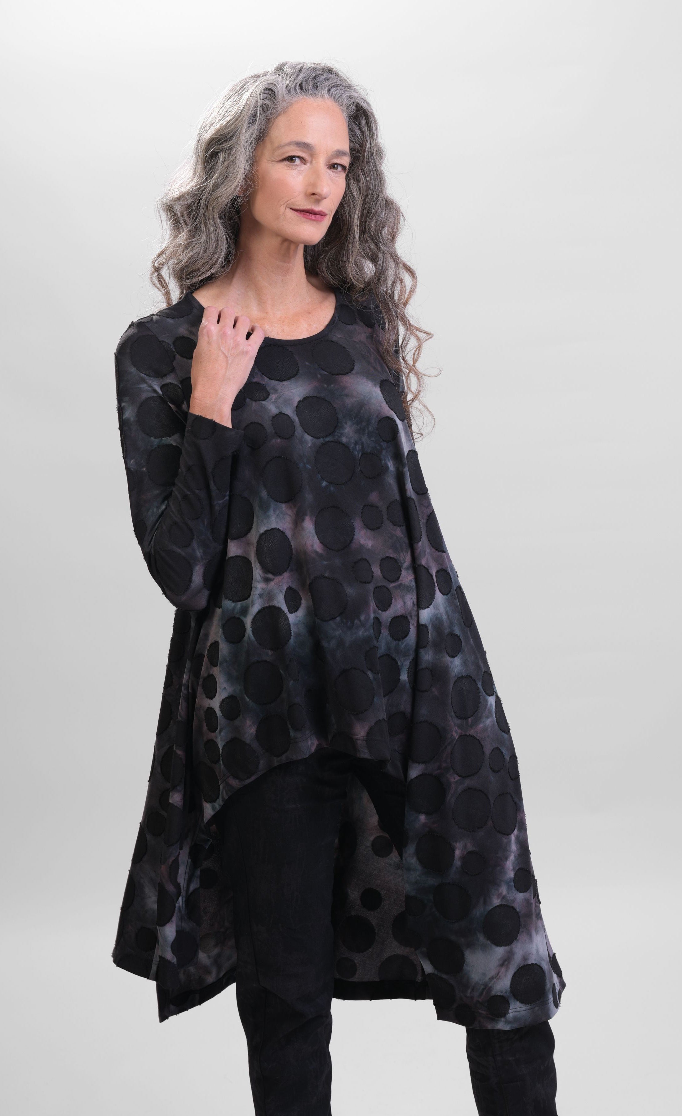 Front top half view of a woman wearing black pants and the alembika smoke echo asymmetric tunic. This tunic is black, blue, and grey tie-dye with black spots all over it. The top has a round neck, long sleeves, and an asymmetrical hi-lo hem.