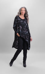 Load image into Gallery viewer, Front full body view of a woman wearing black pants and the alembika smoke echo asymmetric tunic. This tunic is black, blue, and grey tie-dye with black spots all over it. The top has a round neck, long sleeves, and an asymmetrical hi-lo hem.
