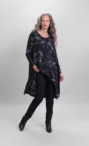 Front full body view of a woman wearing black pants and the alembika smoke echo asymmetric tunic. This tunic is black, blue, and grey tie-dye with black spots all over it. The top has a round neck, long sleeves, and an asymmetrical hi-lo hem.