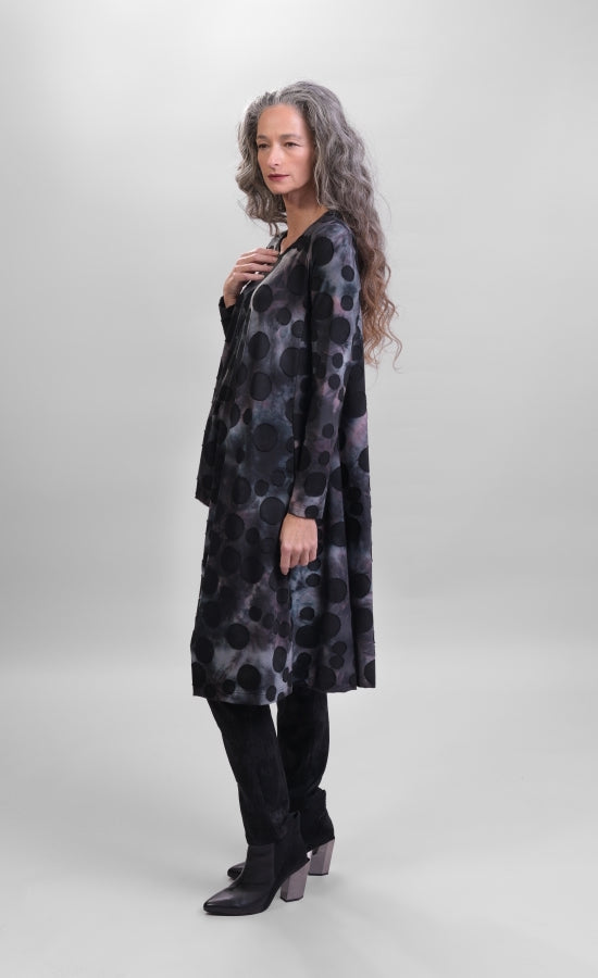 Left side full body view of a woman wearing black pants and the alembika smoke echo asymmetric tunic. This tunic is black, blue, and grey tie-dye with black spots all over it. The top has a round neck, long sleeves, and an asymmetrical hi-lo hem.