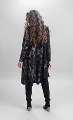 Load image into Gallery viewer, Back full body view of a woman wearing black pants and the alembika smoke echo asymmetric tunic. This tunic is black, blue, and grey tie-dye with black spots all over it. The top has long sleeves and an asymmetrical hi-lo hem.

