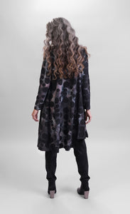 Back full body view of a woman wearing black pants and the alembika smoke echo asymmetric tunic. This tunic is black, blue, and grey tie-dye with black spots all over it. The top has long sleeves and an asymmetrical hi-lo hem.