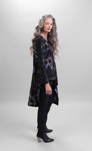 Right side full body view of a woman wearing black pants and the alembika smoke echo asymmetric tunic. This tunic is black, blue, and grey tie-dye with black spots all over it. The top has a round neck, long sleeves, and an asymmetrical hi-lo hem.