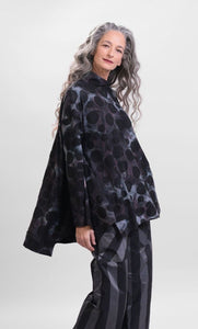 Front top half view of a woman wearing the alembika swing top in smoke. This top is black, grey, and blue tie dye with black dots all over it. The top has a mock neck, long sleeves, and a wide flowy silhouette with a stepped hem.