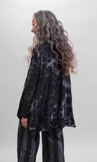 Back top half view of a woman wearing the alembika swing top in smoke. This top is black, grey, and blue tie dye with black dots all over it. The top has a mock neck, long sleeves, and a wide flowy silhouette with a stepped hem.