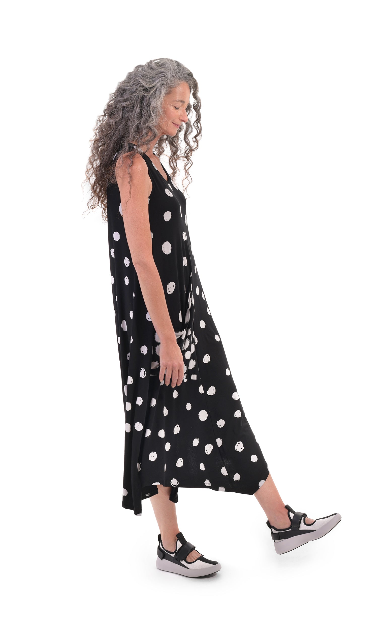 Right sided full body view of a woman wearing the alembika Lia Spotted Jersey Hi-Low Maxi Dress. This sleeveless dress is black with white spiral circles all over it. The front has a scoop neck and a right-sided draped pocket. The hem is asymmetrical and high-low
