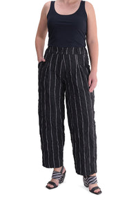 Front bottom half view of a woman wearing the alembika pinstriped pant. This pant is black with white pinstripes. The front has two slanted pockets on the side. The waist is elastic and the pant sits right at the ankles. This pant has a straight leg silhouette.