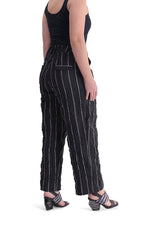 Load image into Gallery viewer, Back, right sided, bottom half view of a woman wearing the alembika pinstriped pant. The pant is black with white pinstripes. The back has two welt pockets. The waist is elastic and the pant sits right at the ankles. This pant has a straight leg silhouette.
