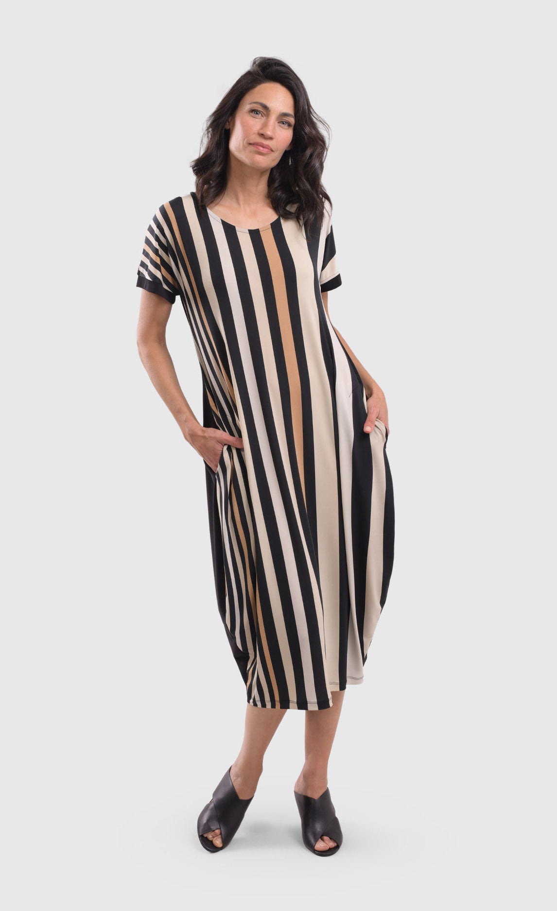 Front full body view of a woman wearing the alembika sunrise stripe dress.  This dress has black and white vertical stripes with a couple of tan stripes in between. The dress has short sleeves, a relaxed silhouette with a poof skirt, and two side pockets. This dress sits mid calf level.