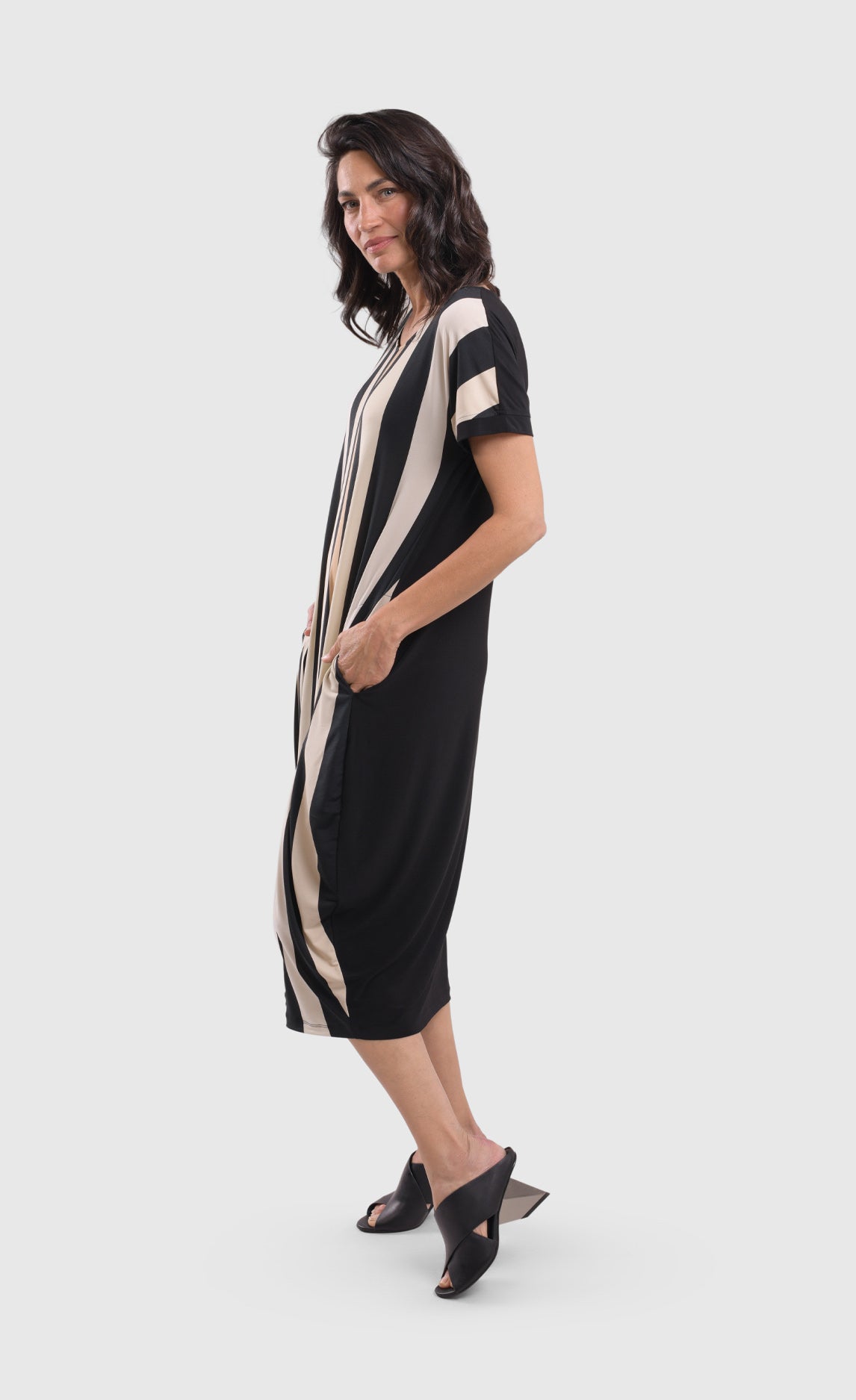 Left side full body view of a woman wearing the alembika sunrise stripe dress. This dress has black and white vertical stripes with a couple of tan stripes in between on the front and a solid back. The dress has short sleeves, a relaxed silhouette with a poof skirt, and two side pockets. This dress sits mid calf level.