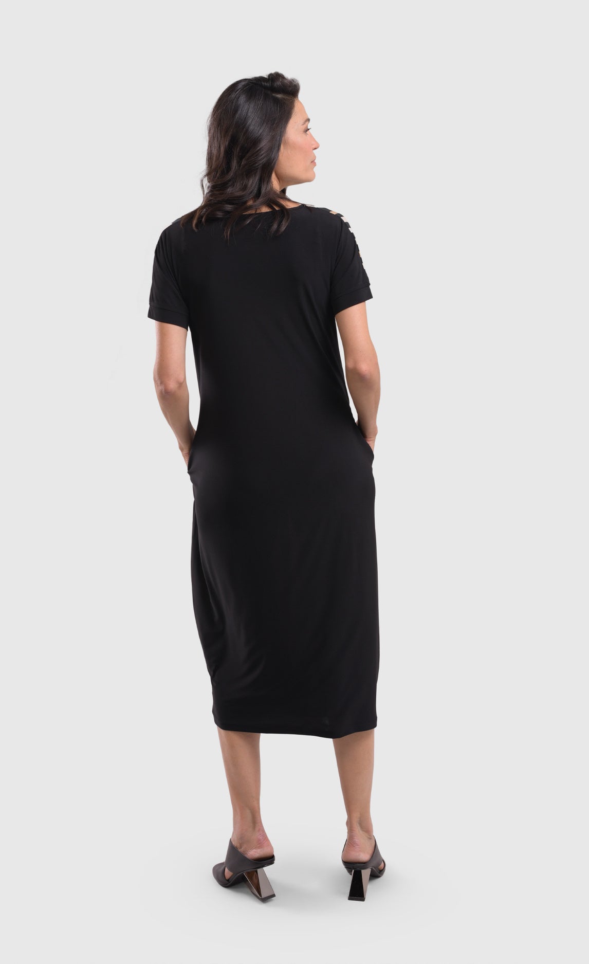 Back full body view of a woman wearing the alembika sunrise stripe dress. This dress is solid black on the back, has short sleeves, and a relaxed silhouette with a poof skirt. This dress sits mid calf level.