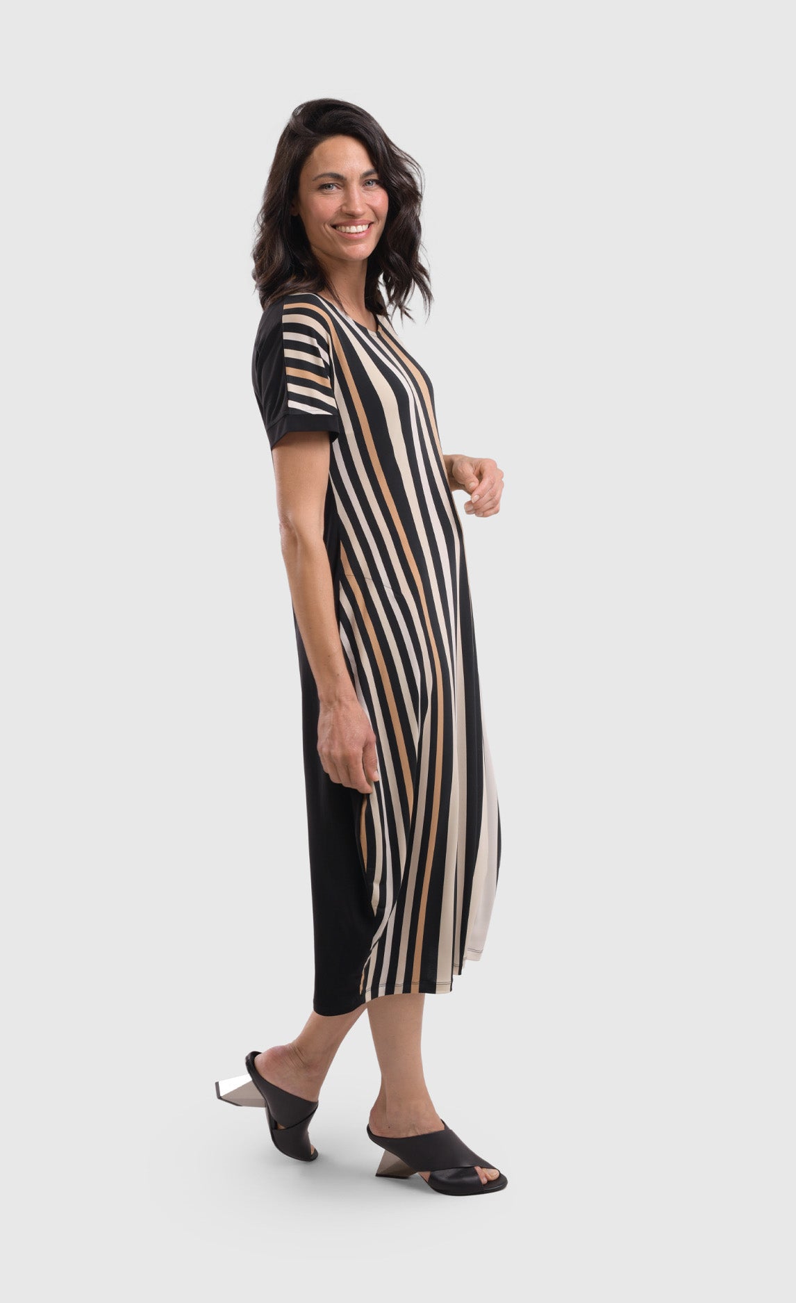 Right side full body view of a woman wearing the alembika sunrise stripe dress. This dress has black and white vertical stripes with a couple of tan stripes in between on the front and a solid back. The dress has short sleeves, a relaxed silhouette with a poof skirt, and two side pockets. This dress sits mid calf level.
