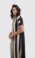 Load image into Gallery viewer, Left side top half view of a woman wearing the alembika sunrise stripe dress. This dress has black and white vertical stripes with a couple of tan stripes in between. The dress has short sleeves, a relaxed silhouette with a poof skirt, and two side pockets. This dress sits mid calf level.
