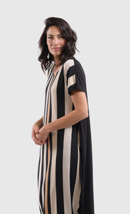 Left side top half view of a woman wearing the alembika sunrise stripe dress. This dress has black and white vertical stripes with a couple of tan stripes in between. The dress has short sleeves, a relaxed silhouette with a poof skirt, and two side pockets. This dress sits mid calf level.