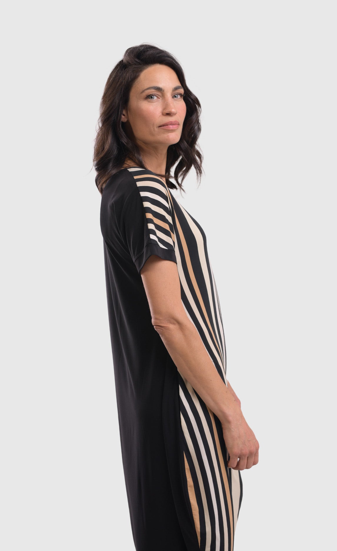 Right side top half view of a woman wearing the alembika sunrise stripe dress. This dress has black and white vertical stripes with a couple of tan stripes in between. The dress has short sleeves, a relaxed silhouette with a poof skirt, and two side pockets. The back of this dress is solid black.