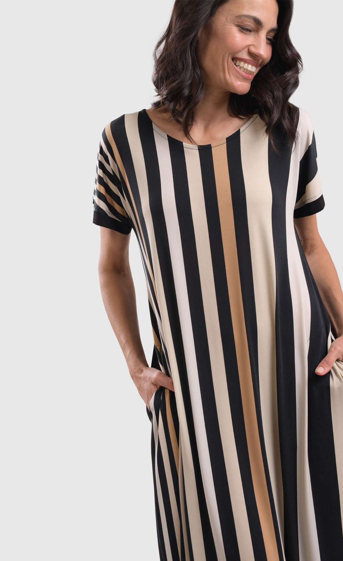 Front close up view of a woman wearing the alembika sunrise stripe dress. This dress has black and white vertical stripes with a couple of tan stripes in between. The dress has short sleeves, a relaxed silhouette with a poof skirt, and two side pockets. 