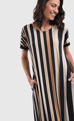 Load image into Gallery viewer, Front close up view of a woman wearing the alembika sunrise stripe dress. This dress has black and white vertical stripes with a couple of tan stripes in between. The dress has short sleeves, a relaxed silhouette with a poof skirt, and two side pockets. 
