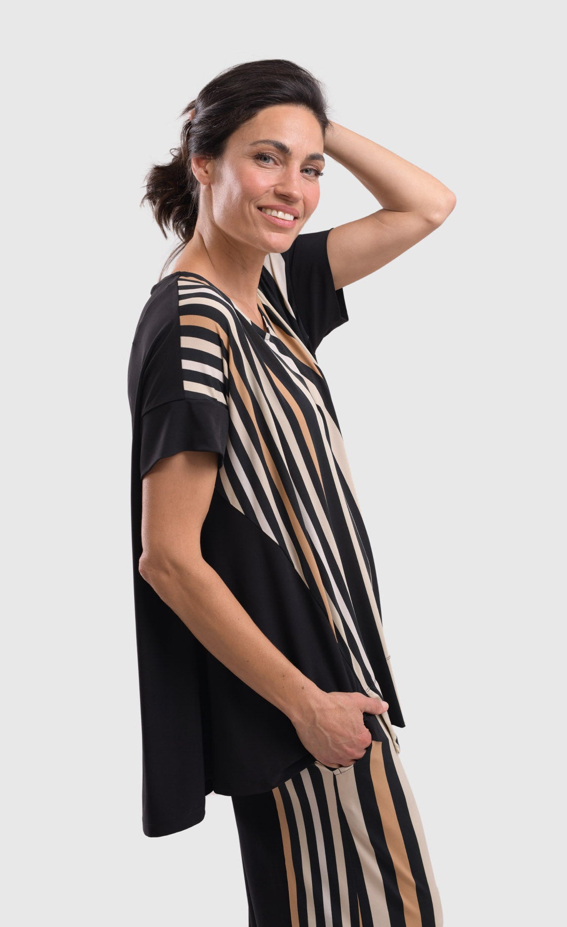Right side top half view of a woman wearing the alembika sunrise stripes swing top. This top has white stripes with a couple of tan stripes in between all of different sizes. The Top has a round neck and short sleeves. the back is solid black.