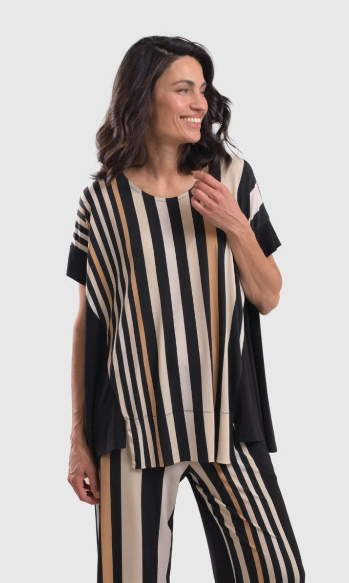 Front top half view of a woman wearing the alembika sunrise stripes swing top. This top has white stripes with a couple of tan stripes in between all of different sizes. The Top has a round neck and short sleeves. the back is solid black.