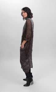 Left side full body view of a woman wearing the alembika talia cocoon russet dress. This dress is slightly sheer with a slip. It has a mix of prints on it that are brown and black and white. The dress has a front draped pocket that can be seen from the left side.