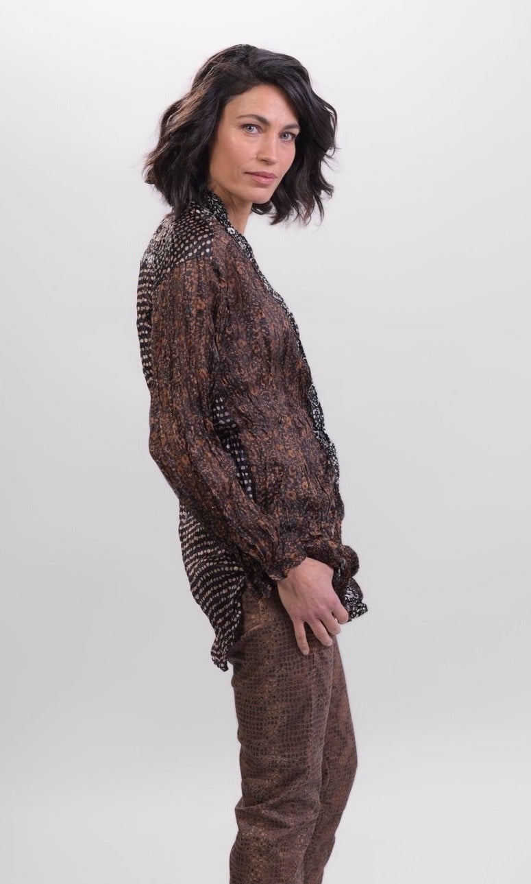 Right side top half view of a woman wearing brown alembika pants and the alembika talia russet blouse. This long sleeved blouse has a button down front, a stand collar, and a mix of different prints that are brown, black, and white.