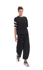 Load image into Gallery viewer, Front full body view of a woman wearing a black tee with stripes on the right arm and the Alembika Riding Pant. This pant is black with wide legs that slightly taper in at the bottom. 

