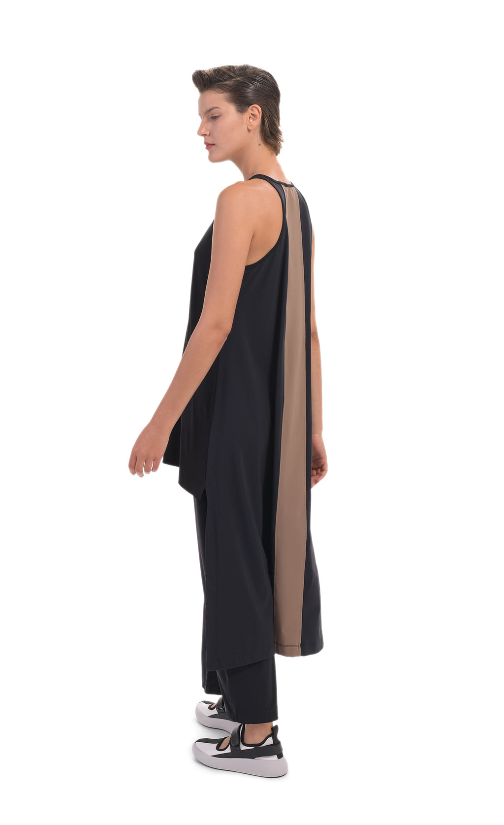 Left, back full body view of a woman wearing the alembika tekbika high low top with the alembika tekbika riding pant. The black top is sleeveless with a t-back. The back of the top has a large brown panel running down the center and comes down to just above the ankles. 