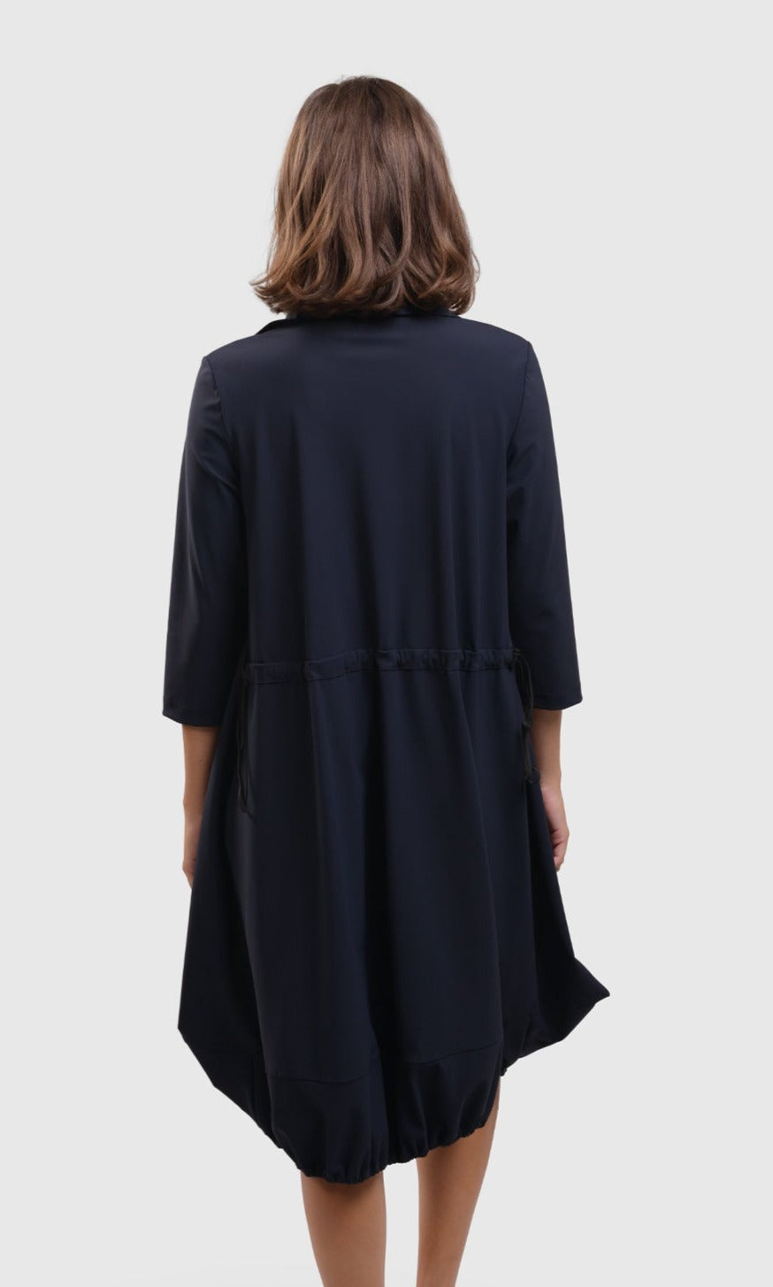 Back top half view of a woman wearing the alembika tekbika navy Dress. This dress sits at the knees and has a bubble skirt and 3/4 length sleeves. The back has a cinch at the waist.