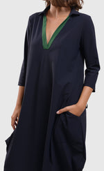Load image into Gallery viewer, Front close up view of a woman wearing the alembika tekbika navy Dress. This dress sits at the knees and has a green v-neck with a collar. The dress has a bubble skirt and 3/4 length sleeves. The woman has her hands in the two front pockets.
