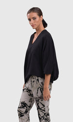 Load image into Gallery viewer, Left side top half view of a woman wearing the alembika urban v neck black top and an alembika grey sketch pant. This top has drop shoulders, an oversized fit, 3/4 length dolman sleeves, and an asymmetrical hem.
