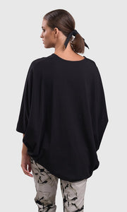 Back top half view of a woman wearing the alembika urban v neck black top and an alembika grey sketch pant. This top has drop shoulders, an oversized fit, 3/4 length dolman sleeves, and an asymmetrical hem.