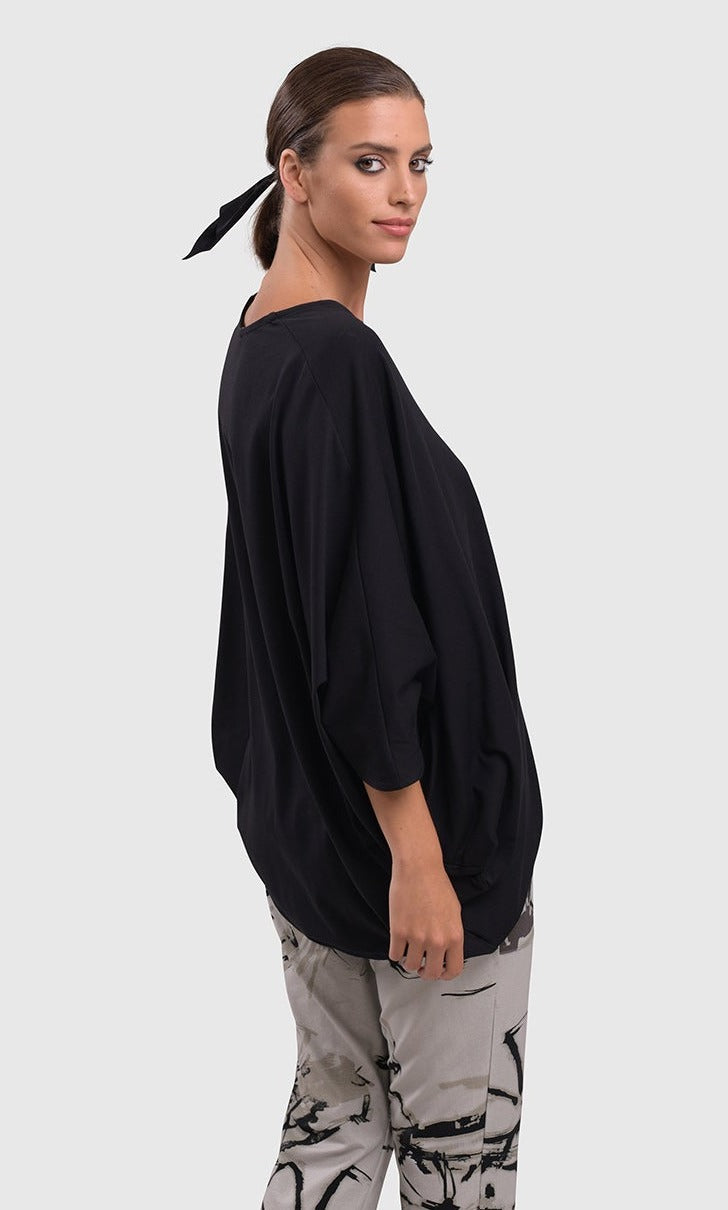 Right side top half view of a woman wearing the alembika urban v neck black top and an alembika grey sketch pant. This top has drop shoulders, an oversized fit, 3/4 length dolman sleeves, and an asymmetrical hem.