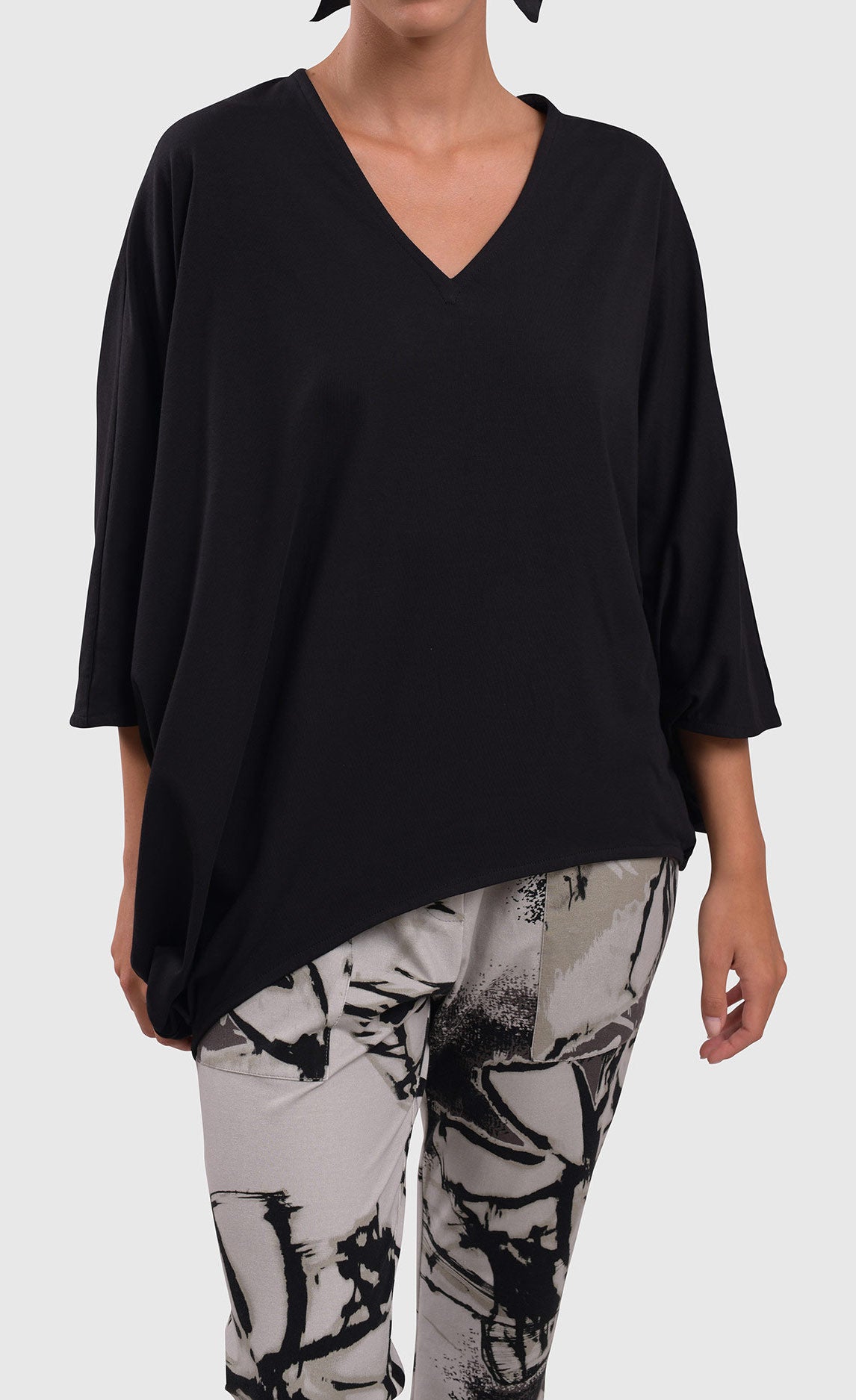 Front top half view of a woman wearing the alembika urban v neck black top and an alembika grey sketch pant. This top has drop shoulders, an oversized fit, 3/4 length dolman sleeves, and an asymmetrical hem.
