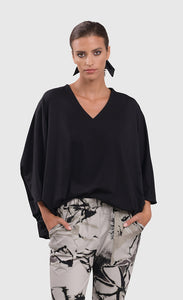 Front top half view of a woman wearing the alembika urban v neck black top and an alembika grey sketch pant. This top has drop shoulders, an oversized fit, and 3/4 length dolman sleeves. 