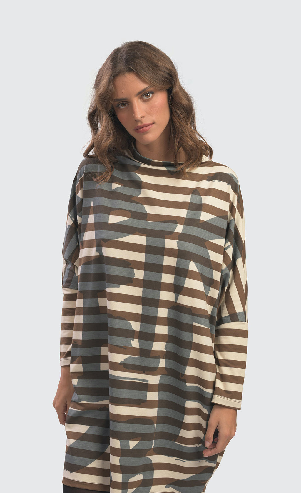 Front top half view of a woman wearing the Alembika Urban Clustur Cocoon Dress. This dress is cream and brown striped with a gray graffiti print all over it. The dress has a cocoon shape that tapers in at the knees. It has drop shoulder, long dolman sleeves and a mock neck.