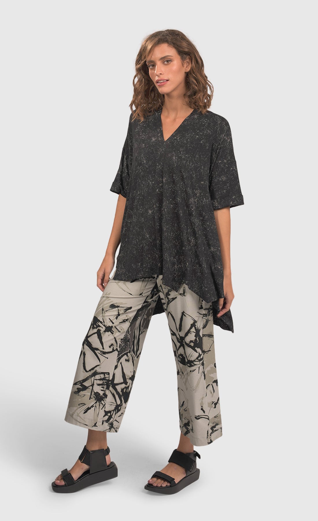 Front full body view of a woman wearing an alembika marble top and the alembika urban sketch go to pant. This pant is grey white with black and dark grey scribbles all over it. The pant has a cropped cut, and wide legs.