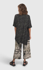 Back full body view of a woman wearing an alembika marble top and the alembika urban sketch go to pant. This pant is grey white with black and dark grey scribbles all over it. The pant has a cropped cut, and wide legs.
