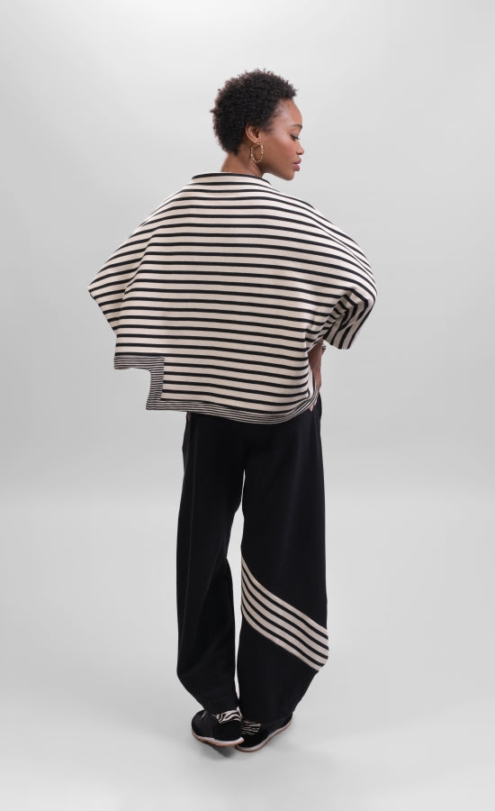 Back full body view of a woman wearing the Alembika Urban French Terry Pants with the Alembika Urban French Terry Stripes Top. This top is white with black striping all over it. The top has an asymmetrical hem, long sleeves, a mock neck, and a boxy silhouette.