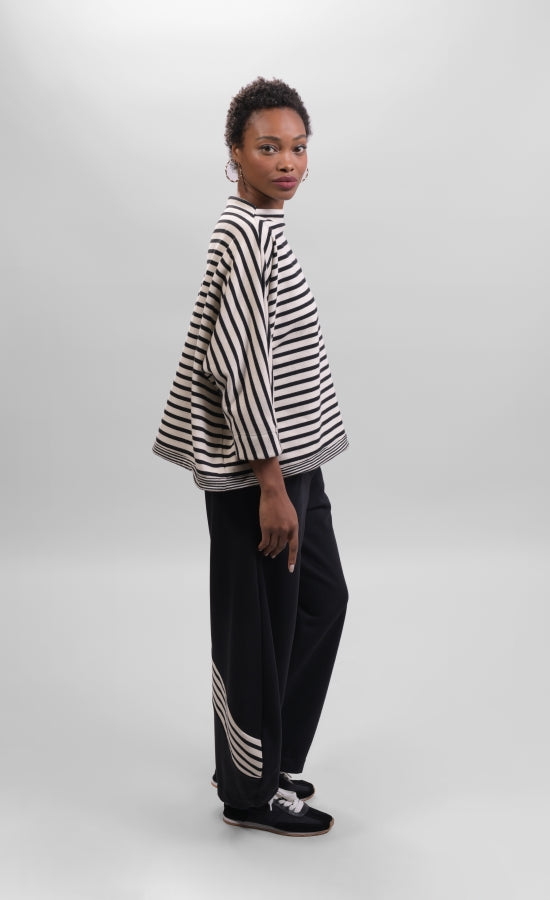 Right side full body view of a woman wearing the Alembika Urban French Terry Pants with the Alembika Urban French Terry Stripes Top. This top is white with black striping all over it. The top has an asymmetrical hem, long sleeves, a mock neck, and a boxy silhouette.