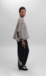 Right side full body view of a woman wearing the Alembika Urban French Terry Pants with the Alembika Urban French Terry Stripes Top. This top is white with black striping all over it. The top has an asymmetrical hem, long sleeves, a mock neck, and a boxy silhouette.
