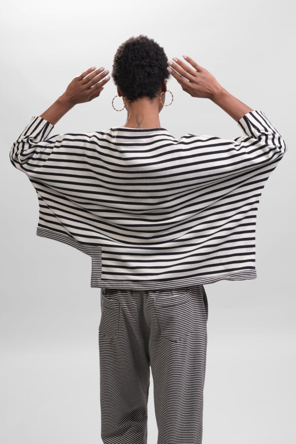 Back top half view of a woman wearing the Alembika Urban French Terry Pants with the Alembika Urban French Terry Stripes Top. This top is white with black striping all over it. The top has an asymmetrical hem, long sleeves, a mock neck, and a boxy silhouette.