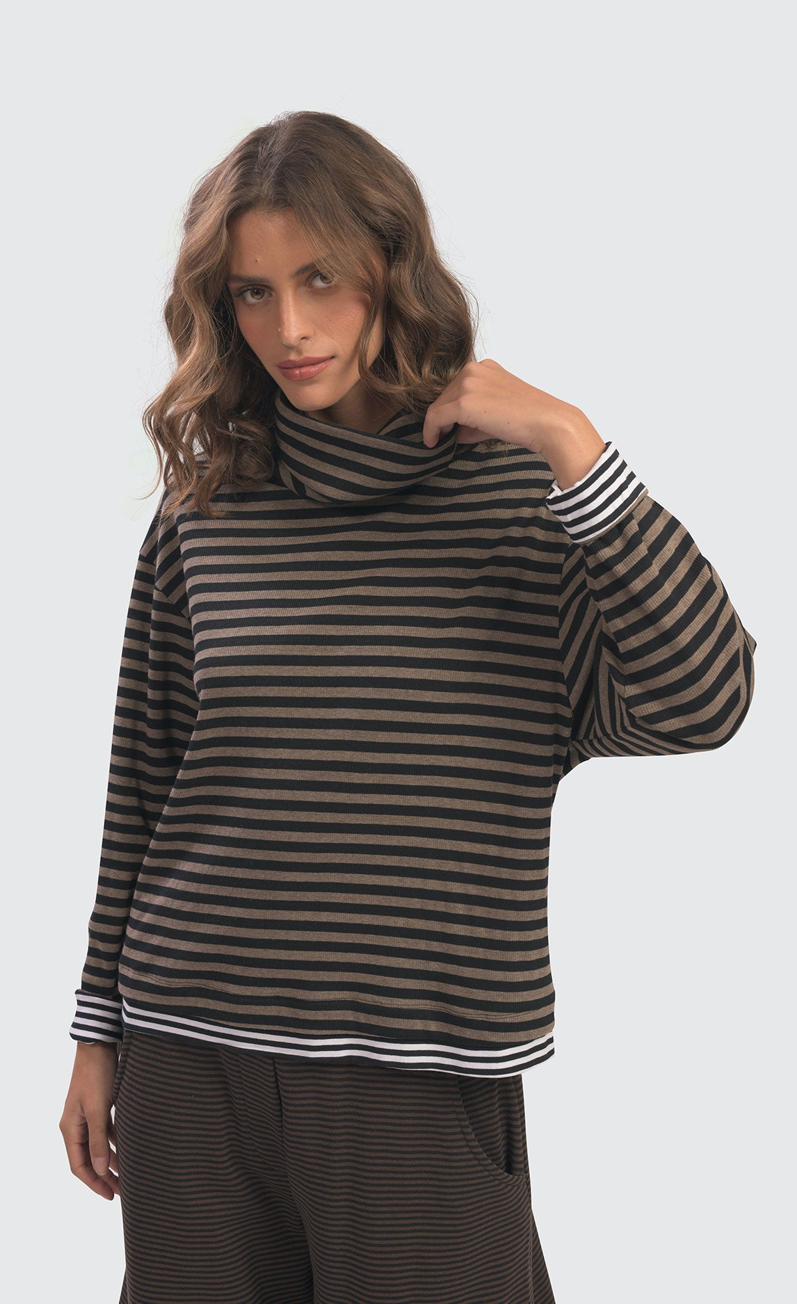 front top half view of a woman wearing the Alembika urban gateau cowlneck top in the brown and black striping. The top has a cowlneck, a relaxed fit, and contrasting black and white hems and cuffs.