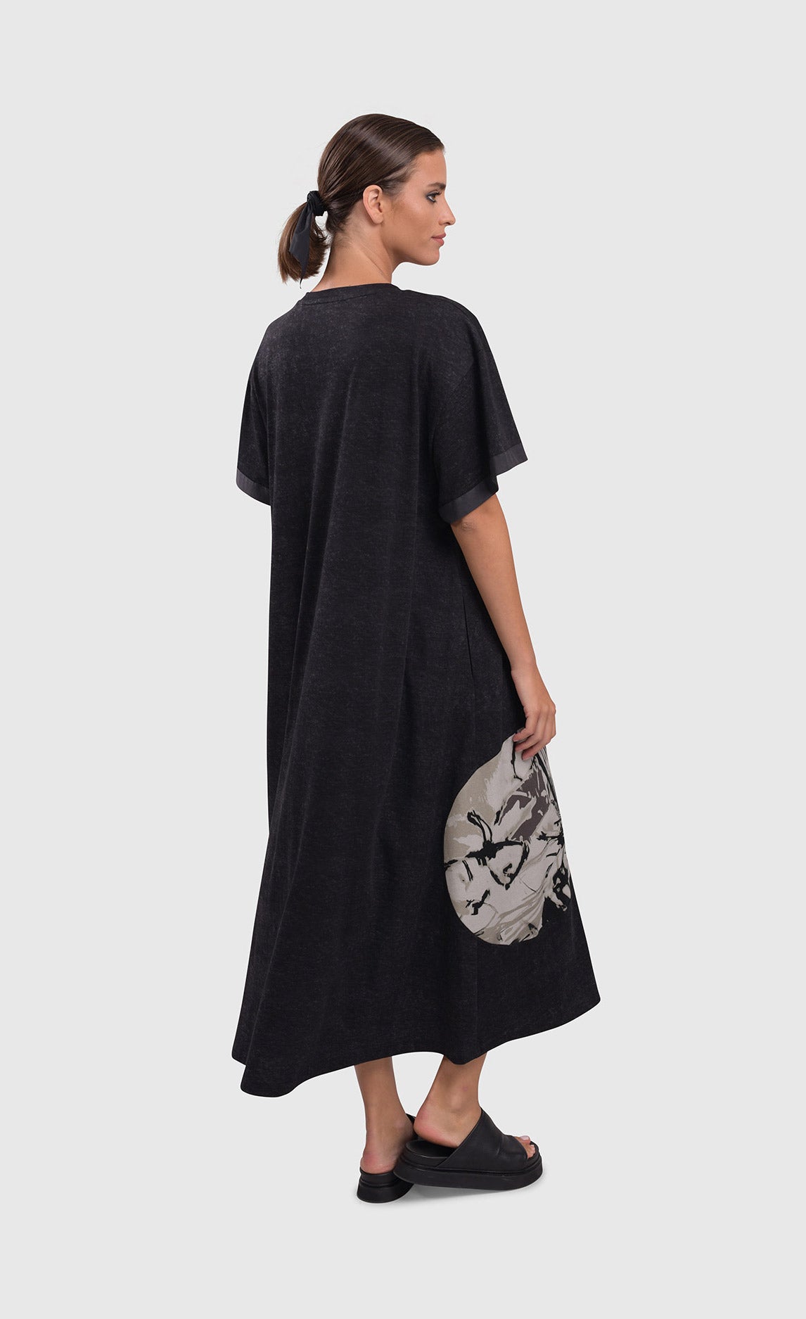 Back full body view of a woman wearing the alembika urban moon maxi dress. This dress is dark grey and a patch circle on the bottom right side of the dress is showing. The circle is white/grey with a scribble print on it. The relaxed dress sits just above the ankles and has elbow length sleeves.