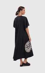 Load image into Gallery viewer, Back full body view of a woman wearing the alembika urban moon maxi dress. This dress is dark grey and a patch circle on the bottom right side of the dress is showing. The circle is white/grey with a scribble print on it. The relaxed dress sits just above the ankles and has elbow length sleeves.
