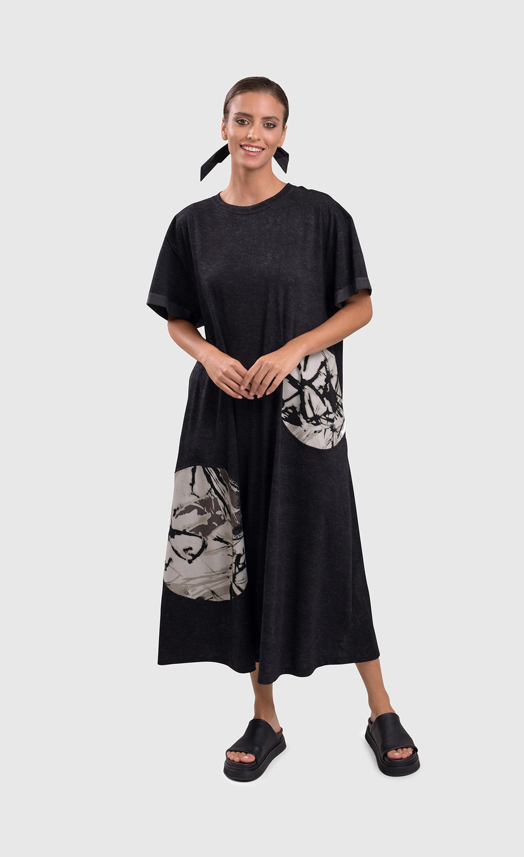 Front full body view of a woman wearing the alembika urban moon maxi dress. This dress is dark grey with two patch circles on the bottom 2/3s of the dress. These circles are white/grey with a scribble print on them. The relaxed dress sits just above the ankles and has elbow length sleeves.