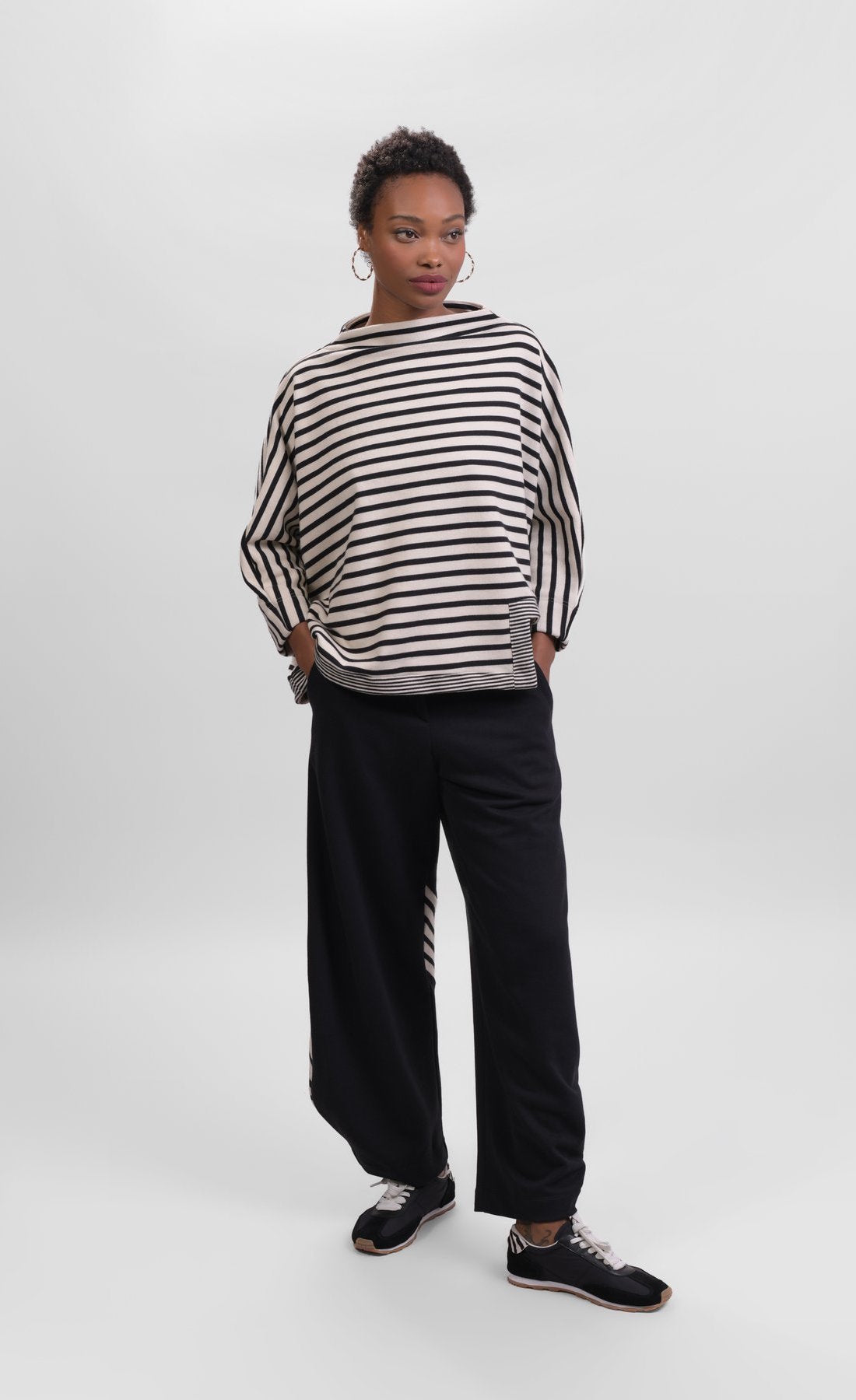Front full body view of a woman wearing the Alembika Urban French Terry Pants with the Alembika Urban French Terry Stripes Top. This top is white with black striping all over it. The top has an asymmetrical hem, long sleeves, a mock neck, and a boxy silhouette.